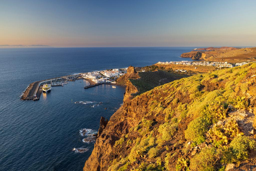 Blick von einer Klippe auf den Hafen von Puerto de las Nieves auf Gran Canaria. Im Vordergrund ragen vulkanische Felsen mit grüner Vegetation ins Bild, unten liegt der Hafen mit einem Fährschiff und weißen Häusern. Dahinter erstreckt sich die Küste bis zum Horizont, umgeben vom blauen Atlantik im warmen Abendlicht.